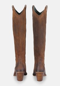 Bottes en daim marron jusqu'au genou avec un design en V, texture lisse et un talon empilé. Le talon présente une légère courbure pour plus de stabilité.