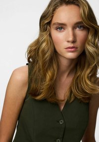 Tamnozelena haljina bez rukava s V-izrezom, s gumbima sprijeda i glatkom teksturom, koju nosi model s valovitom kosom do ramena.