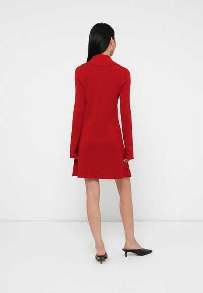 Vrouw met lang zwart haar, gekleed in een lange rode jurk met lange mouwen en zwarte hoge hakken, staand tegen een simpele witte achtergrond, met de rug naar de camera gekeerd.