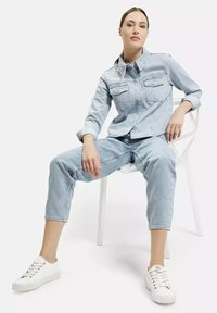 Jasnoniebieska kurtka dżinsowa z dwoma kieszeniami na piersi, w zestawieniu z dopasowanymi jasnoniebieskimi dżinsowymi spodniami 3/4. Model nosi białe sneakersy, siedząc.