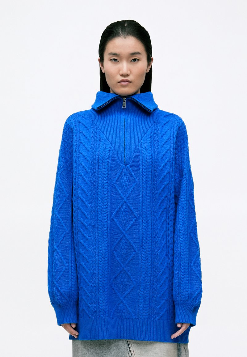Suéter azul de punto con un cuello alto, que presenta patrones de punto trenzado y una cremallera en el cuello. Diseño oversize, mangas largas y tejido texturizado.