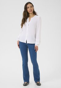 Camicia bianca abbottonata con scollatura rotonda, maniche lunghe e delicati ricami, abbinata a jeans svasati blu e ballerine nere decorate.