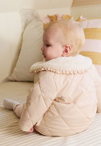 Veste matelassée beige avec un col en fausse fourrure et un détail à volants, portée par un enfant assis sur une surface rayée avec des coussins en arrière-plan.
