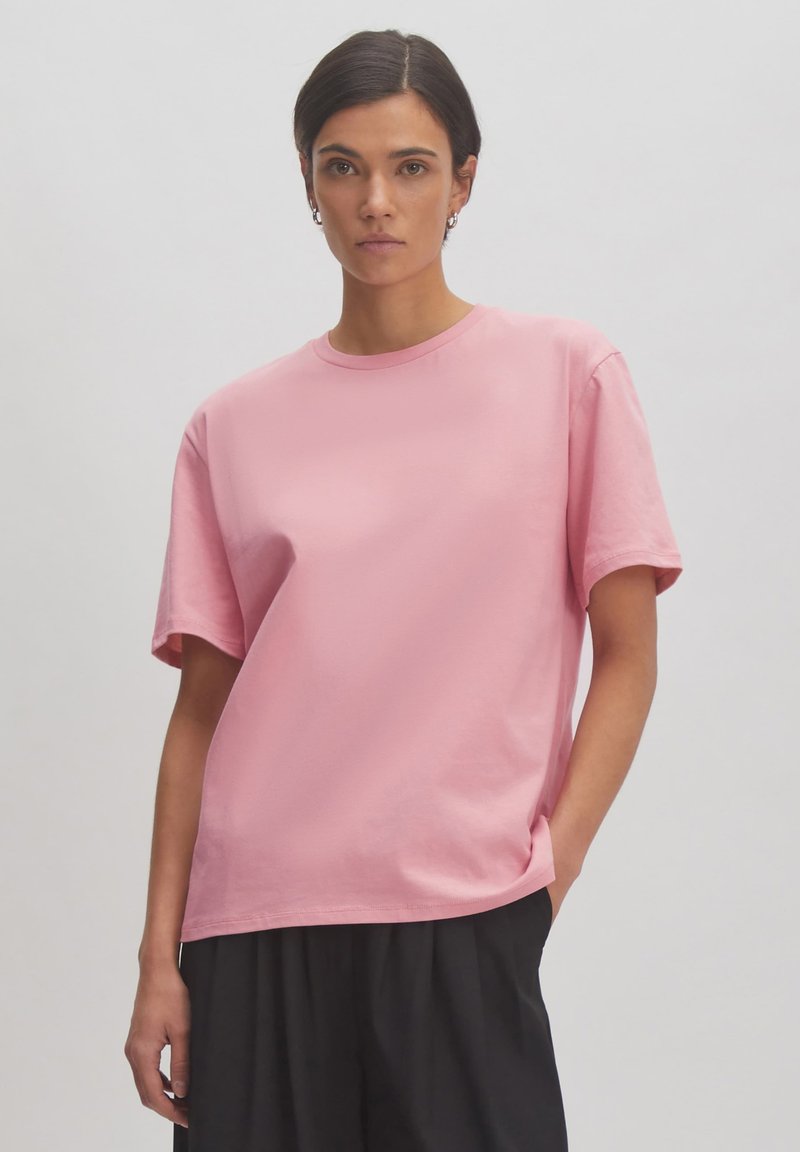 Rosa Baumwoll-T-Shirt mit kurzen Ärmeln und Rundhalsausschnitt, das eine lockere Passform und eine glatte Textur aufweist. Getragen mit weiten schwarzen Hosen.