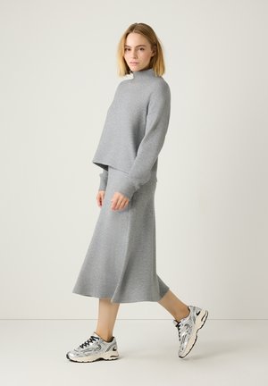 Grauer, kurzer Sweatshirt mit hohem Kragen, kombiniert mit einem passenden, knielangen Rock. Das Modell trägt metallische Sneakers. Glatte Textur.