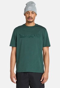 Hombre vestido con una camiseta Timberland de color verde oscuro y un gorro gris claro, de pie contra un fondo blanco liso.