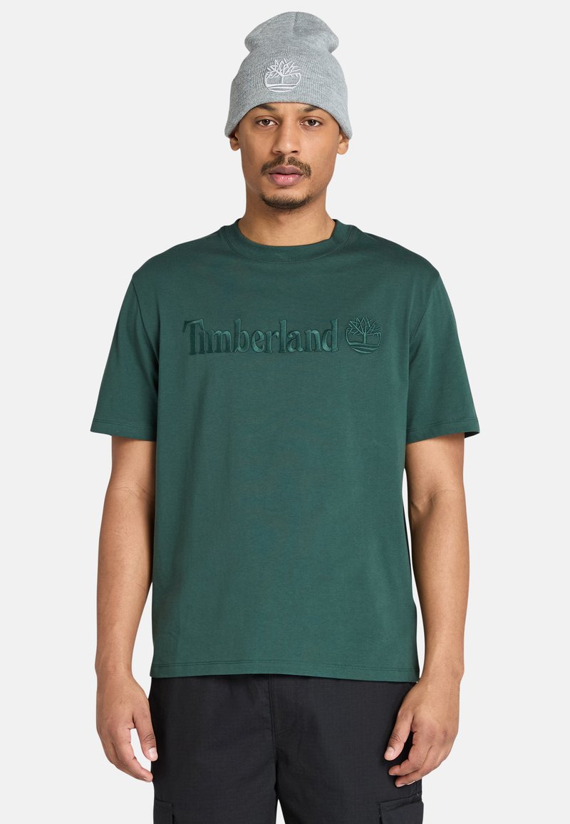 Hombre vestido con una camiseta Timberland de color verde oscuro y un gorro gris claro, de pie contra un fondo blanco liso.