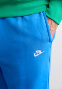 Pantaloni de trening Nike albastri, cu o textură moale, având buzunare laterale și un logo Nike alb deasupra buzunarului drept. Hanoracul verde este vizibil deasupra.