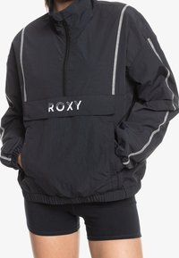 Svart Roxy vindjacka med hög krage, dragkedja fram, elastiska ärmslut och reflektopplight i vitt på ärmar och bröstfickan.