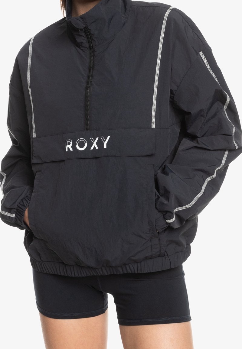 Svart Roxy vindjacka med hög krage, dragkedja fram, elastiska ärmslut och reflektopplight i vitt på ärmar och bröstfickan.