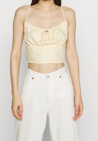 Yellow gingham crop top with a front tie detail, thin straps, and a gathered texture, paired with white high-waisted jeans.