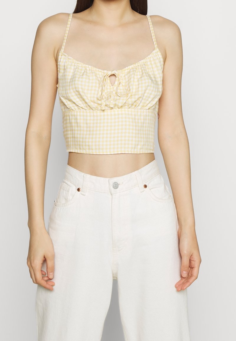 Yellow gingham crop top with a front tie detail, thin straps, and a gathered texture, paired with white high-waisted jeans.