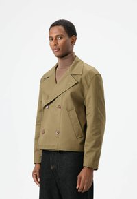 Olijfgroene dubbele rij jas met grote bruine knopen, ingezette revers en zijzakken. Draagt over een bruine coltrui.