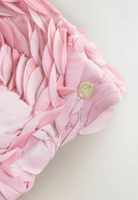 Ružová textilná čelenka s vrstvenými detailmi v tvare lupienkov a malým okrúhlym zlatým príveskom vyrytým s písmenom "B".