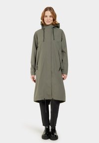 Impermeable largo color verde oliva con capucha, cordones ajustables, dos bolsillos laterales y un corte holgado. Botines negros hasta el tobillo usados debajo.