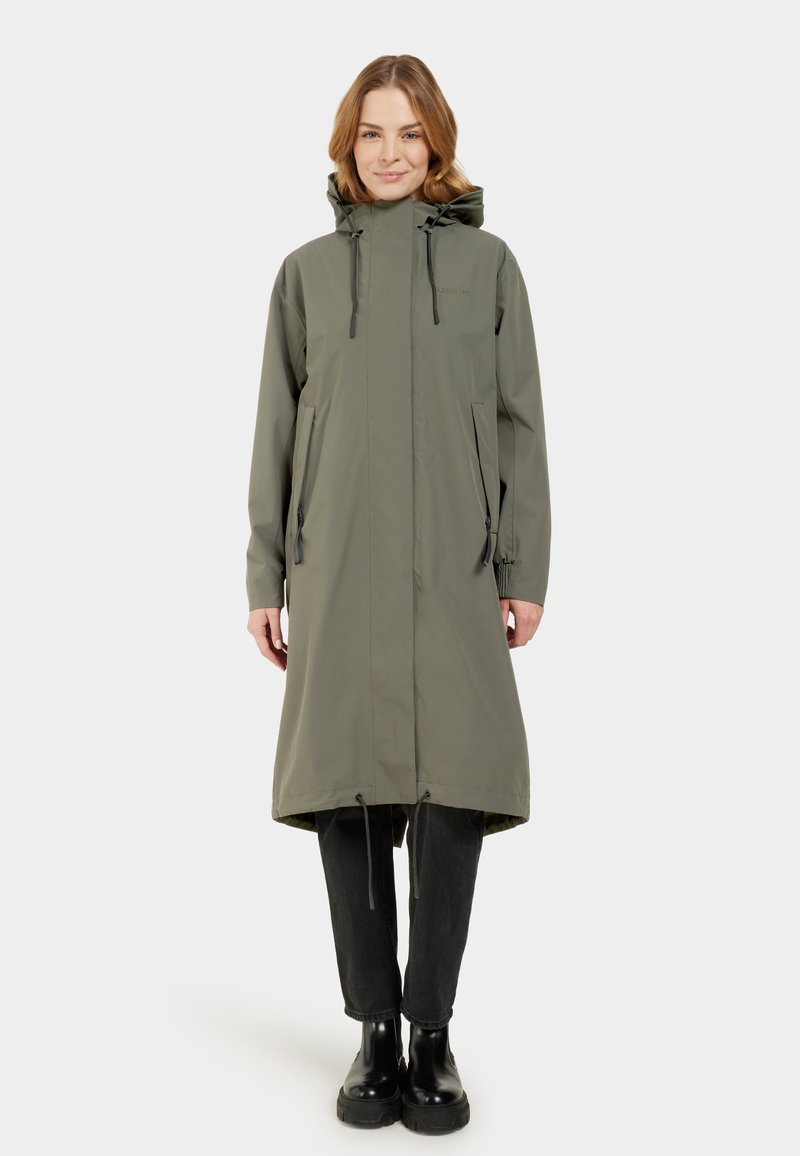 Impermeable largo color verde oliva con capucha, cordones ajustables, dos bolsillos laterales y un corte holgado. Botines negros hasta el tobillo usados debajo.