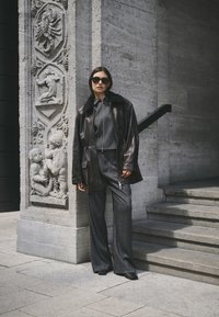 Zwarte leren jas met bontkraag, grijs textuur tweedelig outfit en zwarte schoenen, staande naast een versierde stenen muur en trap.