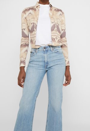 Persoon draagt lichtblauwe hoogwater flared jeans, wit T-shirt en beige patroon blouse met lange mouwen.
