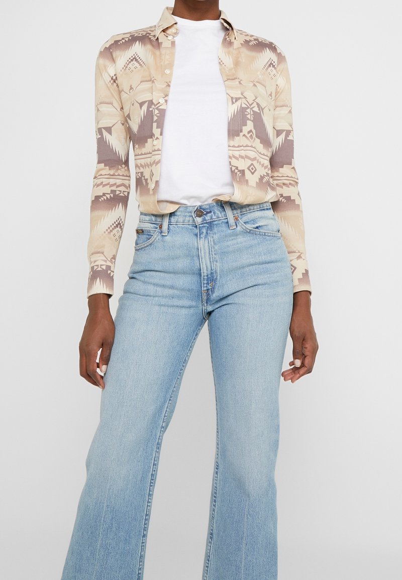 Personne portant un jean évasé taille haute bleu clair, un t-shirt blanc et une chemise à motifs beige à manches longues avec boutons.