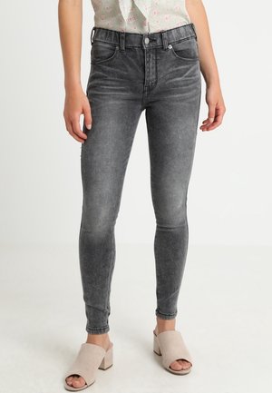 Femme portant un jean slim noir délavé, un chemisier blanc à fleurs et des sandales beiges à talon bloc et bout ouvert, debout devant un fond uni.