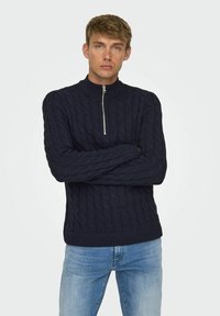 Marineblauer Strickpullover mit hohem Kragen und halbem Reißverschluss, ausgestattet mit gerippten Bündchen und Saum. Die Textur ist erhaben und zeigt ein klassisches Muster.