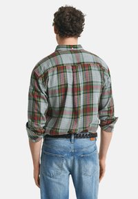 Geruite shirt in groen, rood en wit met gemengde patronen, lange mouwen opgerold tot aan de ellebogen, en een rugplooi. Draagt met lichtblauwe jeans.
