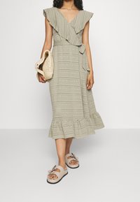 Khaki plisovaná šaty s V-výstřihem a rukávy s nabíráním, s očkovým detailem a volánkovým lemem. V kombinaci s béžovými skládacími sandály a tkanou taškou.