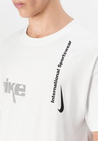 Hvid bomulds t-shirt med sort trykt tekst "Nike" og "International Sportswear" samt et swoosh logo, rund halsudskæring og løs pasform.
