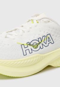 Baskets de sport blanches avec tige en maille, logo HOKA proéminent en bleu, accents jaunes et semelle intermédiaire texturée. Bout arrondi et semelle rembourrée.