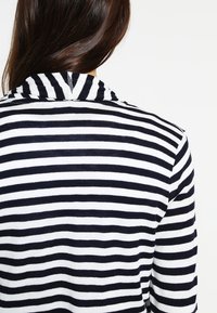 Persona con cabello largo castaño, vistiendo una camiseta de rayas horizontales en blanco y negro, vista desde atrás contra un fondo blanco.