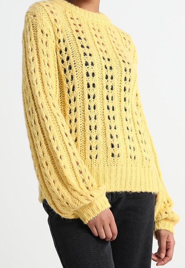 Pull à grosse maille jaune avec des motifs ajourés verticaux, manches longues et poignets côtelés, porté avec un pantalon noir.