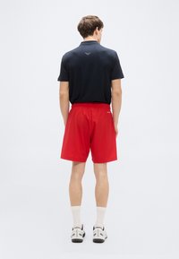 Μαύρο πόλο μπλουζάκι με λογότυπο στην πλάτη, συνδυασμένο με κόκκινα σορτς. Το μοντέλο φοράει λευκές κάλτσες και μαύρα αθλητικά παπούτσια, στέκεται σε απλό φόντο.