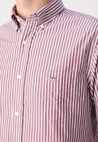Gros plan d'une personne portant une chemise à rayures verticales rouges et blanches avec une poche poitrine brodée d'un petit logo.