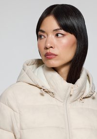 Chaqueta acolchada beige con cierre frontal, capucha ajustable y detalles de costura en tono. Textura suave y diseño esbelto.