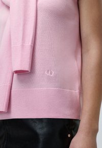 Lys rosa strikket genser med ribbestrikket hem og mansjetter, med en liten brodert logo detalj nær bunnen.
