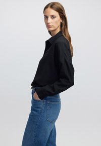 Schwarzes Langarmshirt mit strukturiertem Kragen und lockerer Passform, kombiniert mit hellblauen Denim-Jeans. Der Stoff wirkt glatt mit subtiler Textur.