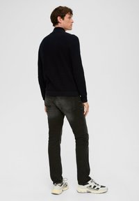 Czarny sweter z dzianiny z wysokim kołnierzem, połączony z ciemnymi dżinsami. Model w masywnych białych i czarnych sneakerach, widoczny z tyłu.