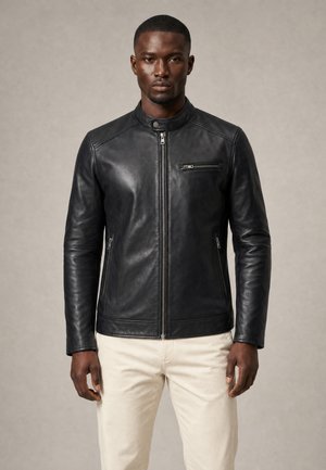Mann trägt schwarze, lederne Jacke mit Reißverschluss und Stehkragen sowie hellbeige Hose, steht vor neutralem Hintergrund.
