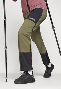 Pantalons de randonnée verts et noirs avec poches zippées, fabriqués en matériau durable. Comprend des accents texturés noirs au niveau de l'ourlet et un détail de logo.