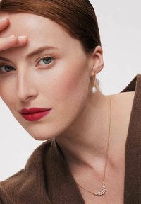 Złoty naszyjnik z małymi cyrkoniami w kształcie półksiężyca. Modelka nosi kolczyki z perłowym zapięciem, które ładnie komponują się z gładką fakturą brązowego stroju.