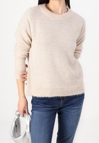 Hellbeige gestrickter Pullover mit Rundhalsausschnitt und gerippten Bündchen, der eine weiche Textur aufweist. Kombiniert mit dunkelblauen Jeans und einer silbernen Handtasche.