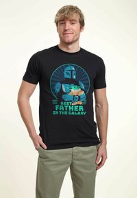 Black t-shirt with graphic of a character in armor holding a creature, text reads "Best Father in the Galaxy," featuring blue and green accents.