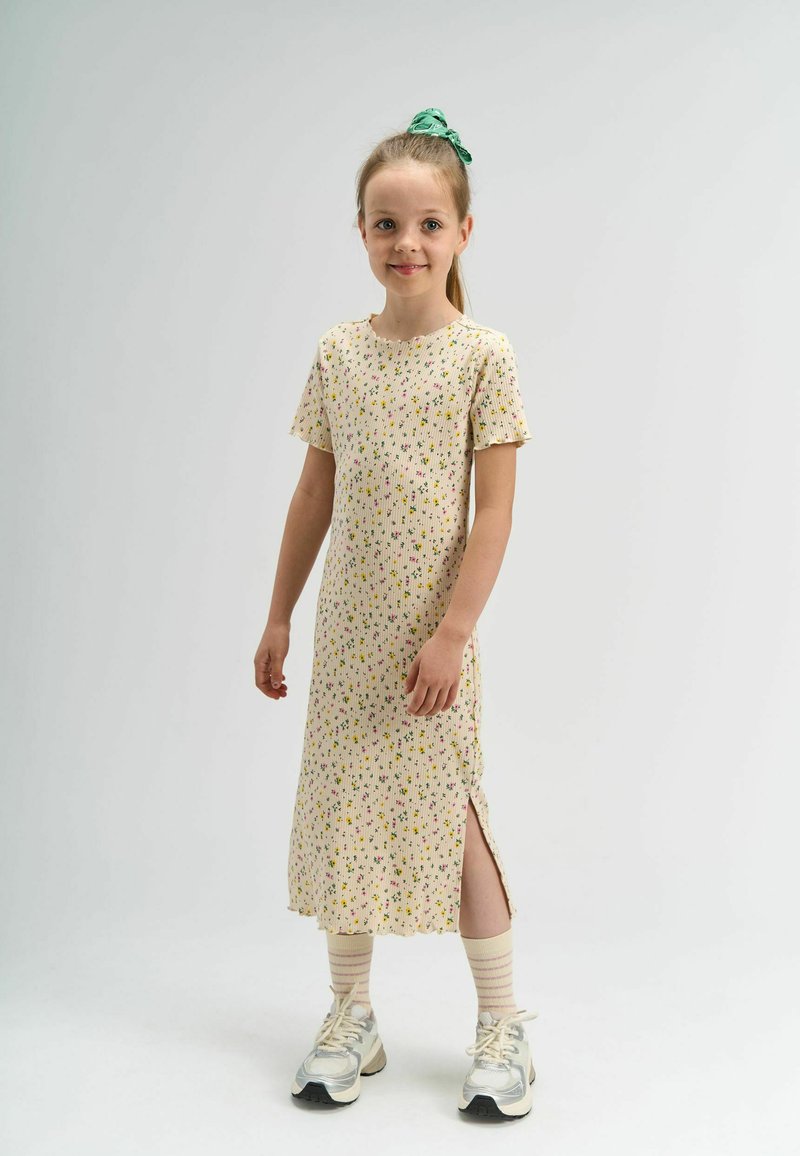 Jeune fille souriante, portant une robe crème mi-longue avec un petit motif floral, des chaussettes rayées légères, des baskets argentées et un chouchou vert.