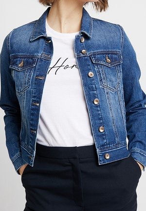 Vrouw draagt een blauwe spijkerjas over een wit shirt met de tekst "Home" en zwarte broek, handen in de zakken.