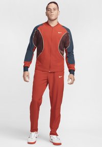 Trening sportiv roșu, cu accente bleumarin și maronii, jachetă cu fermoar, pantaloni ajustați și detaliu cu logo. Fabricat dintr-un material neted și ușor.