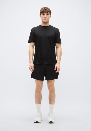 Μαύρο αθλητικό μπλουζάκι και σορτς, εφαρμοστή σχεδίαση, λεία υφή. Το μοντέλο φοράει λευκές κάλτσες και αθλητικά παπούτσια, στέκεται μπροστά σε απλό φόντο.