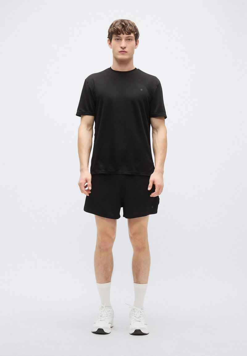 Μαύρο αθλητικό μπλουζάκι και σορτς, εφαρμοστή σχεδίαση, λεία υφή. Το μοντέλο φοράει λευκές κάλτσες και αθλητικά παπούτσια, στέκεται μπροστά σε απλό φόντο.