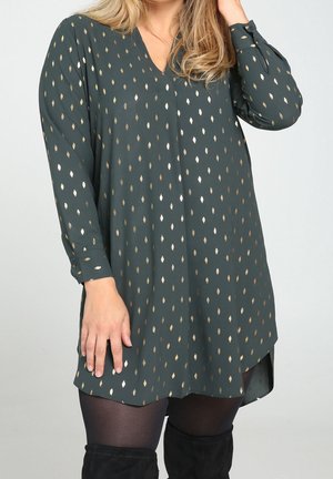 Green tunic with a V-neck, long sleeves, and gold leaf pattern. Soft texture, rounded hem, and buttoned cuffs. Worn over black tights.