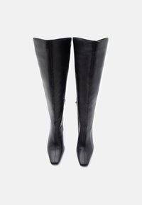 Bottes en cuir noir montantes au-dessus du genou avec un design à bout carré. Caractérisées par une texture lisse et des fermetures éclair latérales pour faciliter l'enfilage.