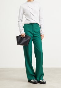 Bílý pletený svetr s spirálovým vzorem, zkombinovaný s vysokými zelenými kalhotami. Černá taška s zakřiveným vrchem a boty s kovovými detaily.